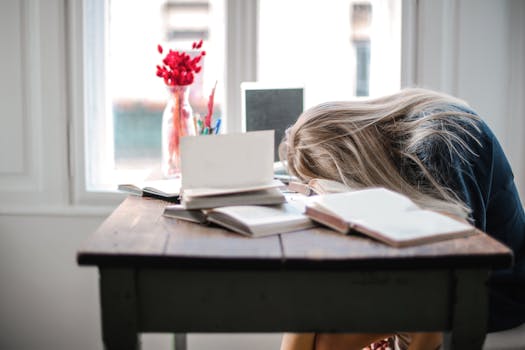 Blonde woman slumped over desk filled with books, showcasing study fatigue indoors.