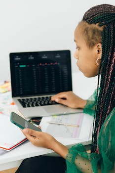 Focused woman working with financial data on laptop and smartphone in modern office.