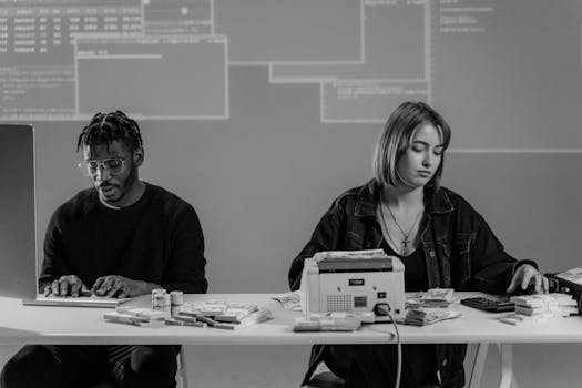 Two professionals working with computers and currency at a desk in a modern office setting.